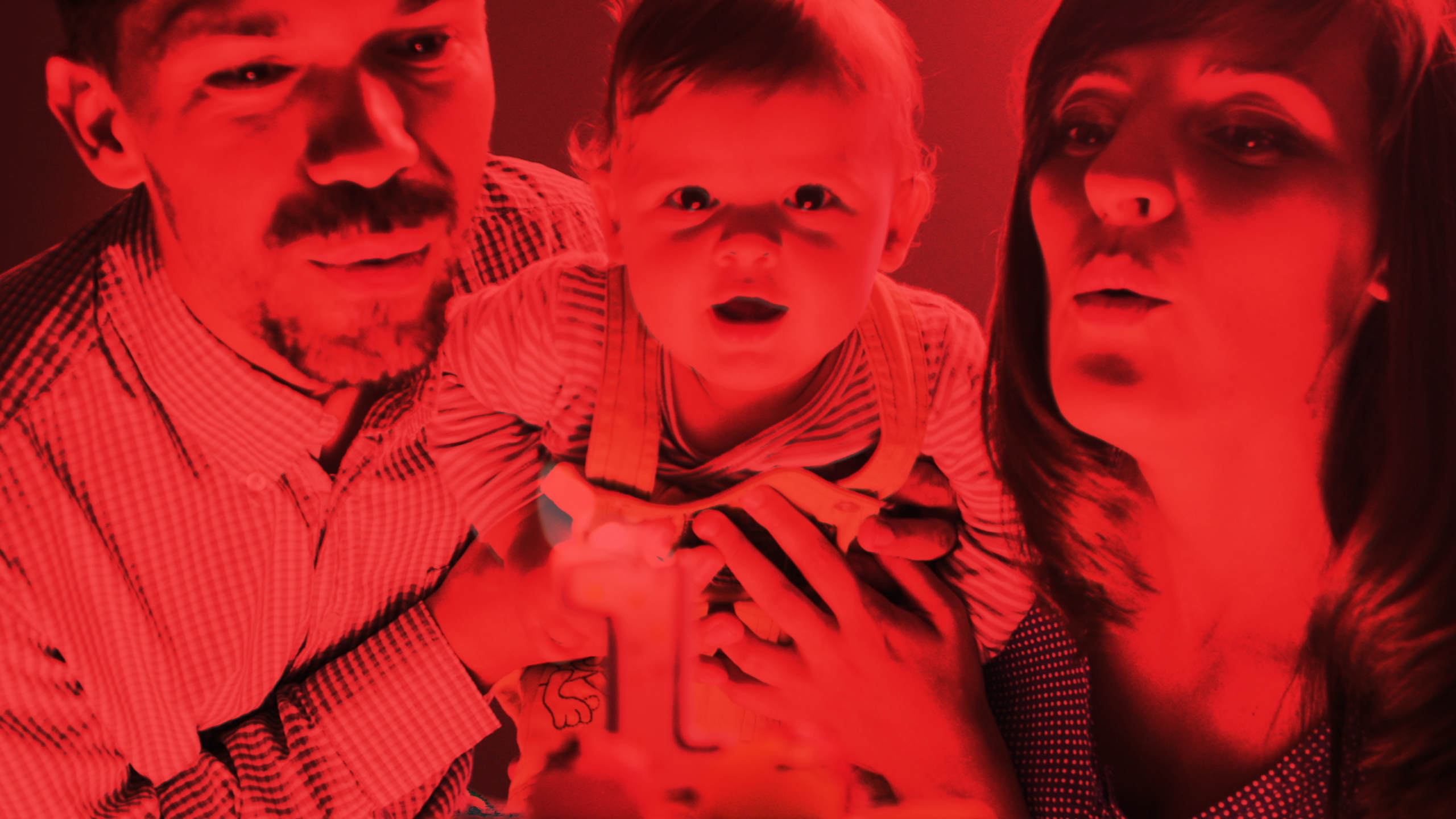 Image tinted red, a man and woman hold their infant. Together, they blow out the one-shaped birthday candle.